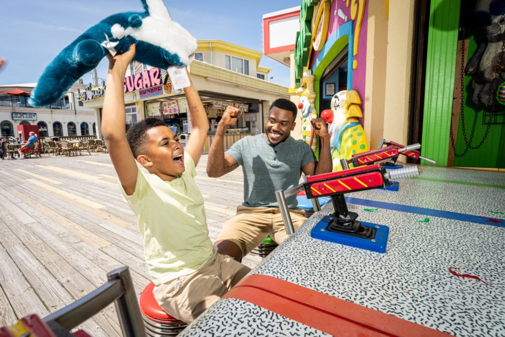 A father and son enjoy water gun game at Jenkinson's Boardwalk. Son holds up stuff shark toy after winning game