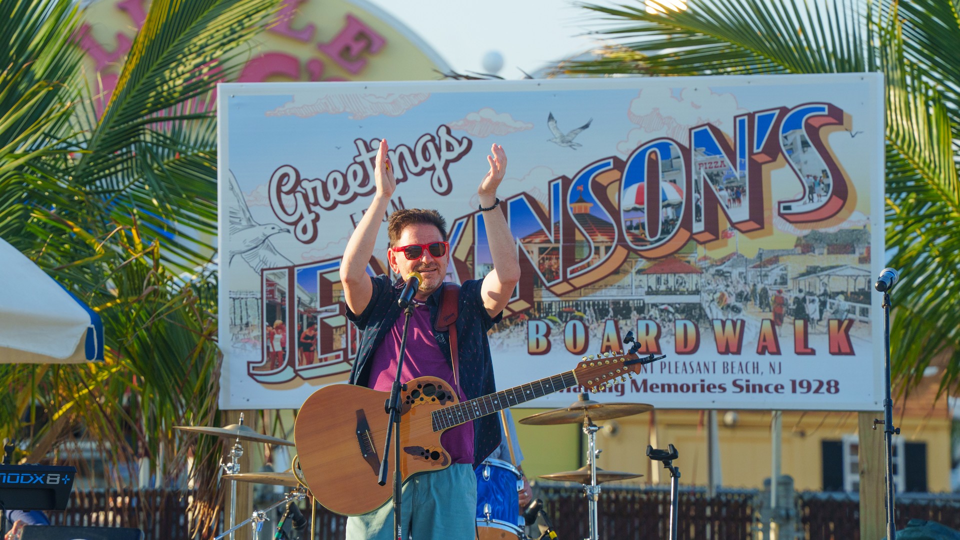 Musician performs under a large "Greensons Boardwalk" sign outdoors on a sunny day.