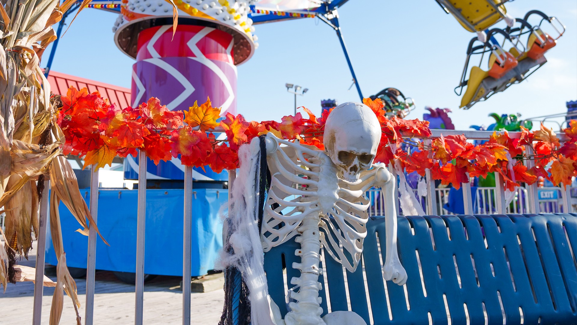 A skeleton decoration sits on a bench with fall garland and rides in the background at Jenkinson’s Boardwalk.