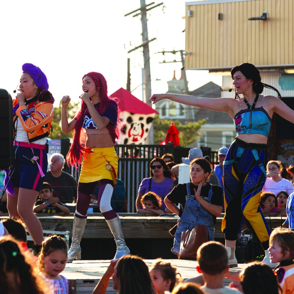 Performers in colorful costumes sing and dance during the Popstar Sing-A-Long as children and families gather around the stage at Jenkinson's Boardwalk.
